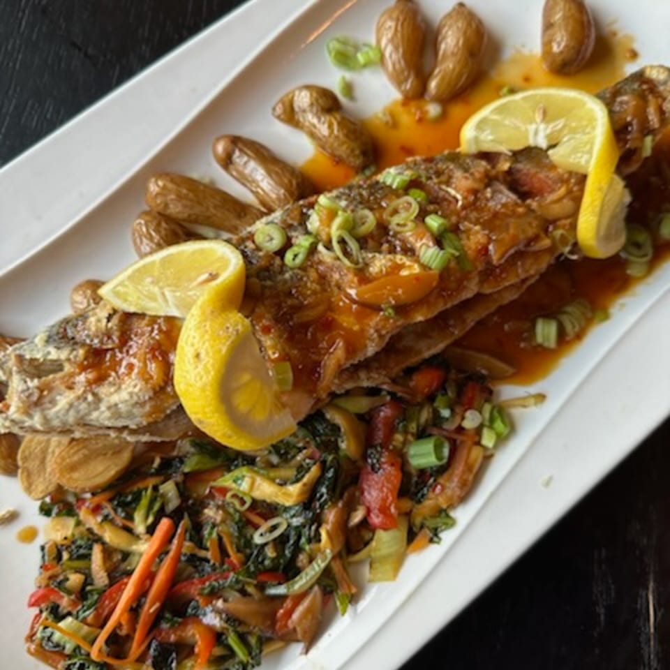 Whole fried fish with lemon slices, roasted potatoes, and stir-fried vegetables on a white platter.