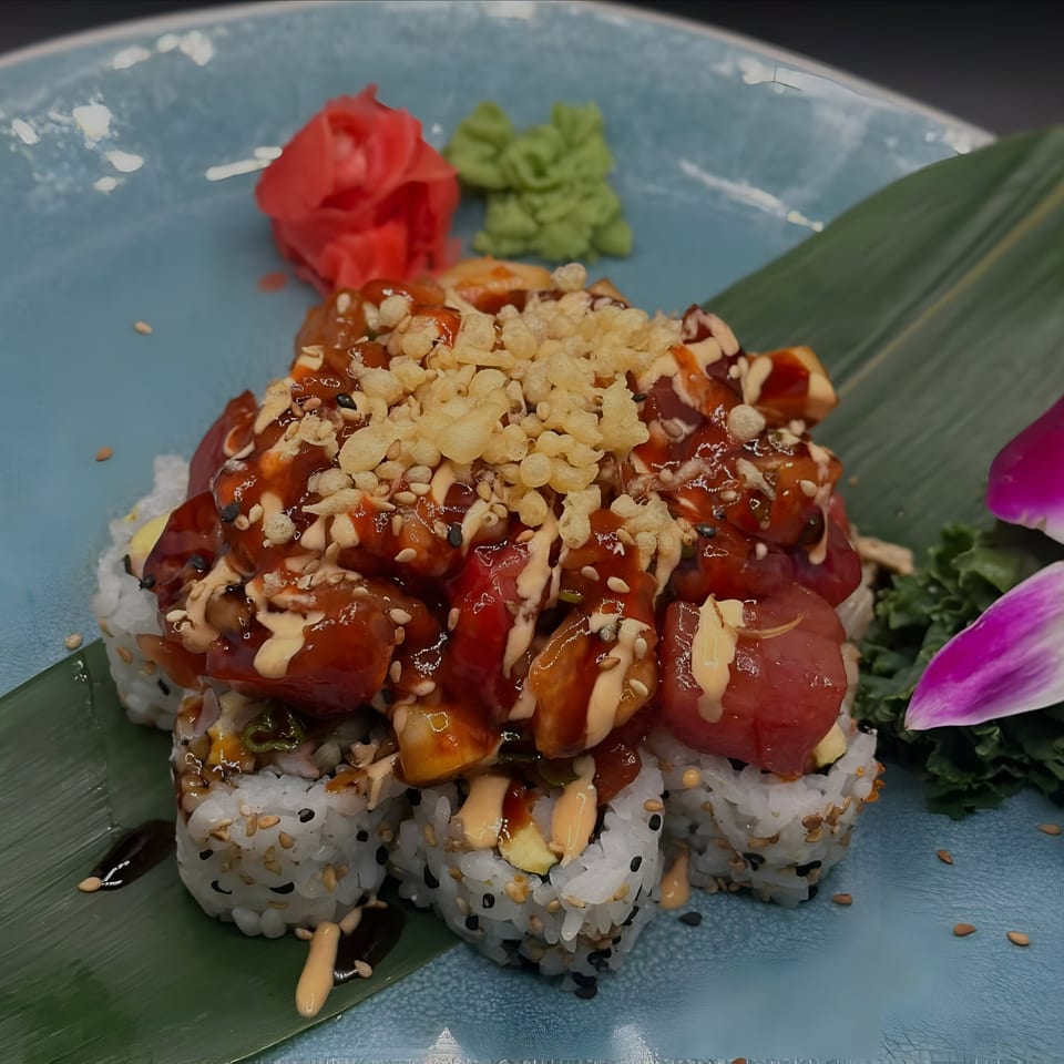 Sushi rolls topped with tuna, sauce, and crispy bits, garnished with ginger, wasabi, and an orchid.