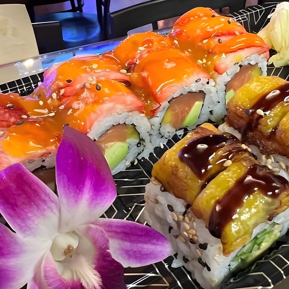 Assorted sushi rolls with vibrant orange and brown sauces, garnished with a purple orchid.