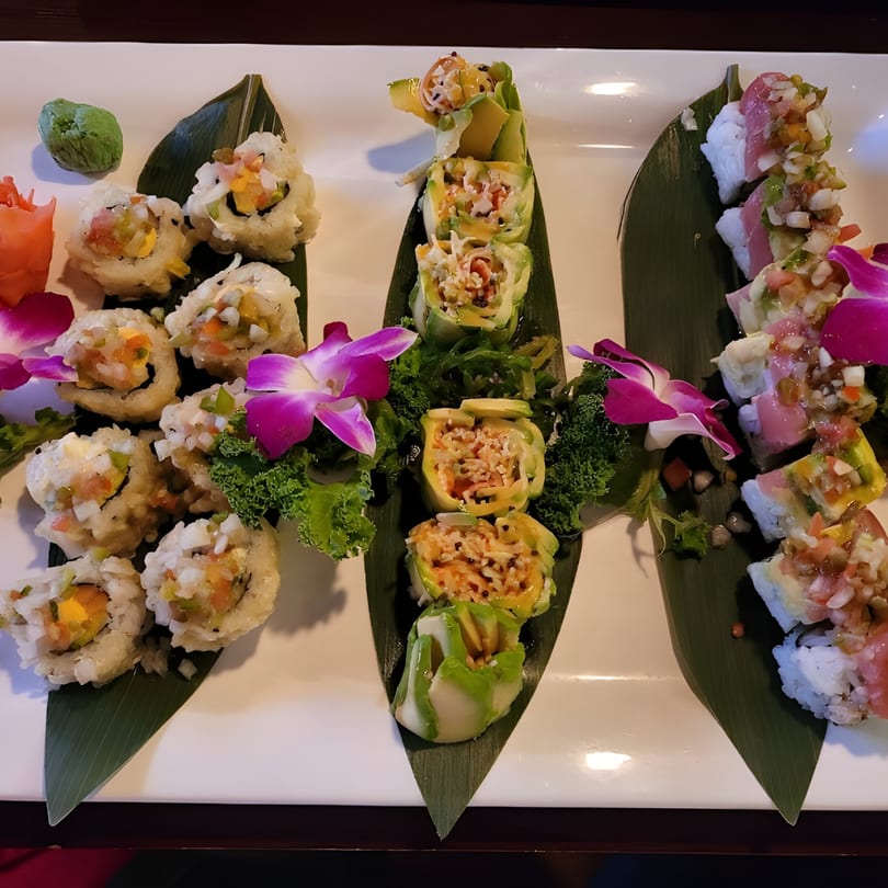 Three rows of colorful sushi rolls and sashimi, garnished with orchids and kale, on a white platter.