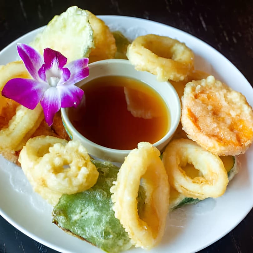 Tempura platter with various fried vegetables and calamari rings, served with a dipping sauce and orchid.
